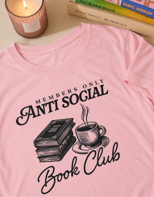 Pink t-shirt with 'Anti Social Book Club' design featuring books and a cup on a table with a candle and books.