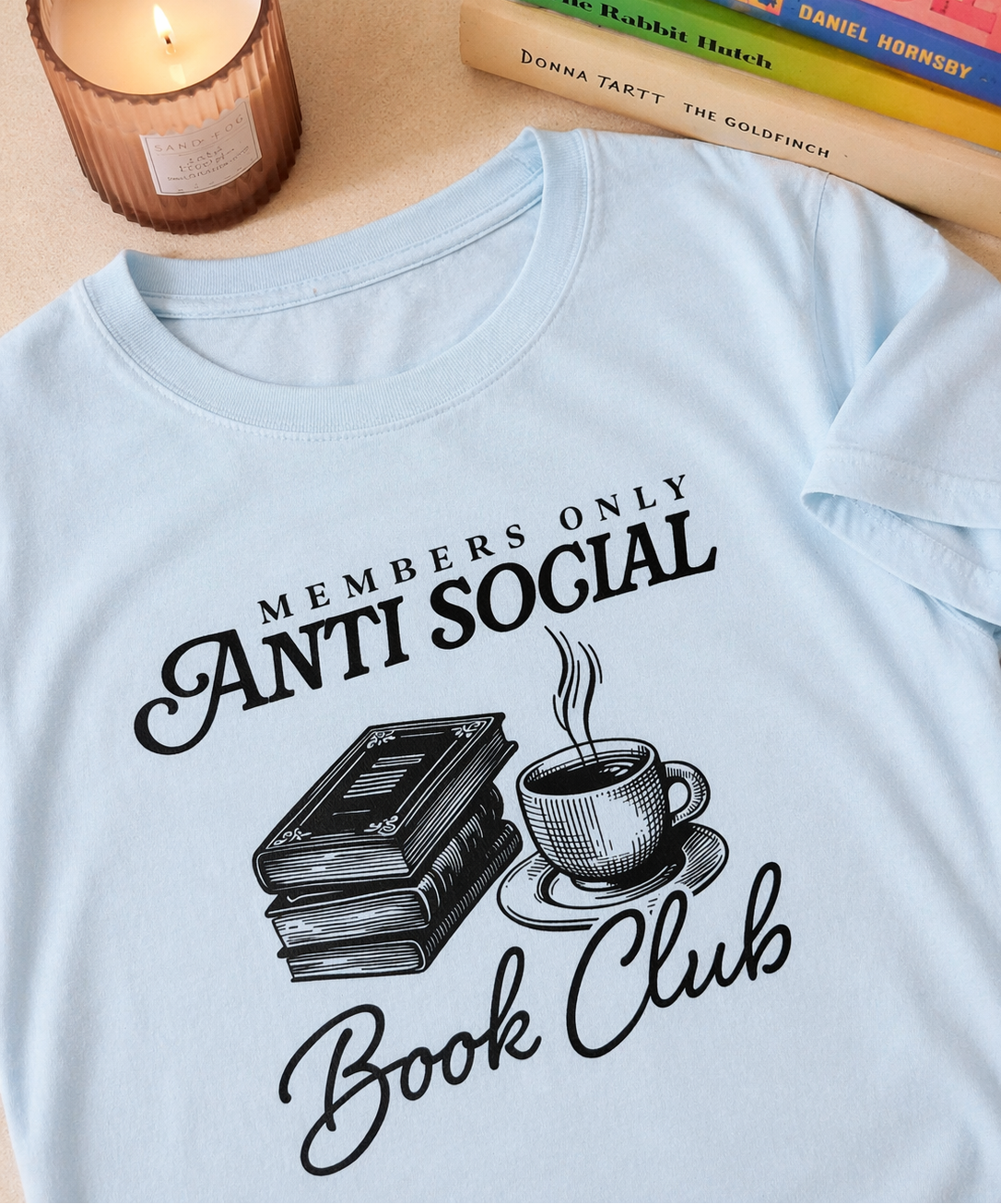 Light blue t-shirt with 'Members Only Anti Social Book Club' design featuring books and a coffee cup, placed on a surface with books and a candle.
