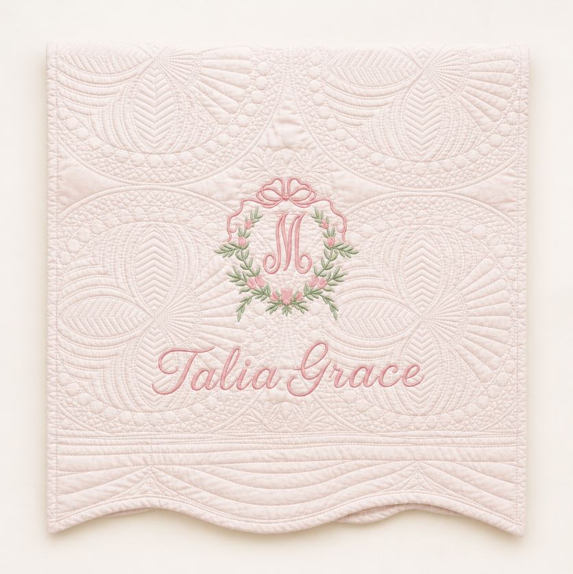 A light pink quilted fabric panel with scalloped edges, featuring an embroidered monogram 'M' in a floral wreath and the name 'Talia Grace' in pink script.