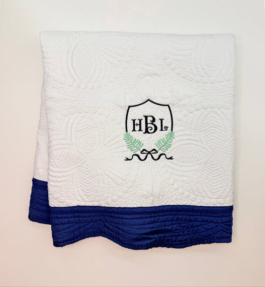 A folded white quilted blanket with a navy blue bottom border and a custom embroidered crest featuring the initials 'HBL' in black, green leaves, and a bow, displayed against a light beige background.