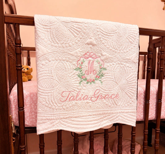 Baby crib with a white quilt featuring floral embroidery and embroidered name, placed on a wooden crib.