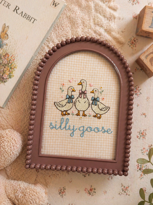 embroidered silly goose nursery frame with beaded mauve arch frame styled on soft neutral flatlay with books and baby decor