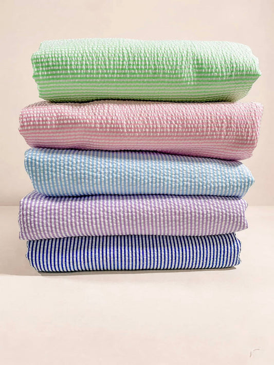 A stack of five folded textured cotton fabrics in pastel colors, including mint green, pink, light blue, lavender, and navy blue, set against a warm beige background.