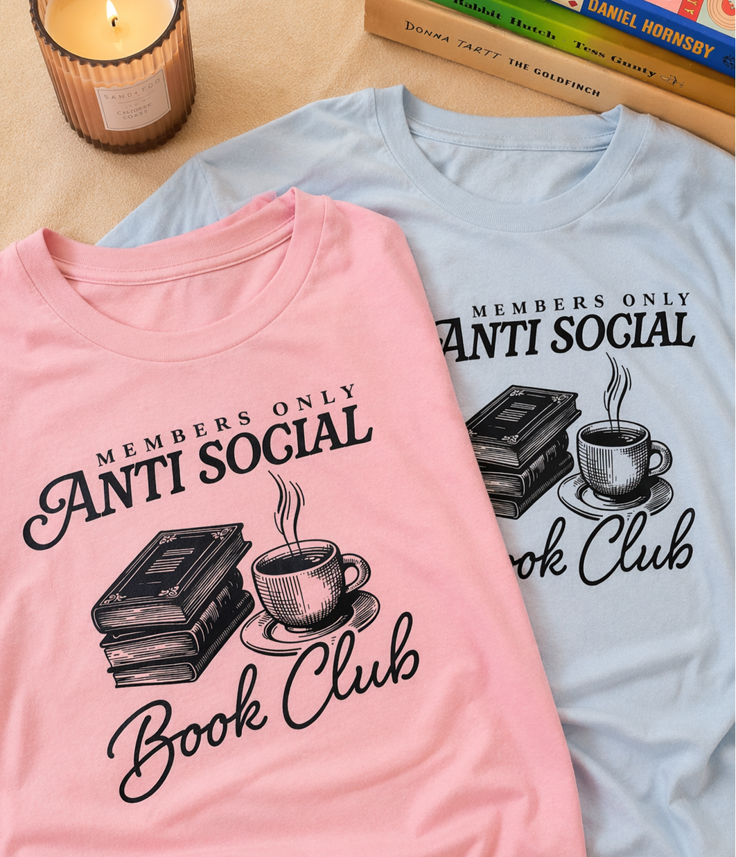 Pink and light blue graphic t-shirts with “Members Only Antisocial Book Club” design featuring books and coffee, styled with candle and stacked books