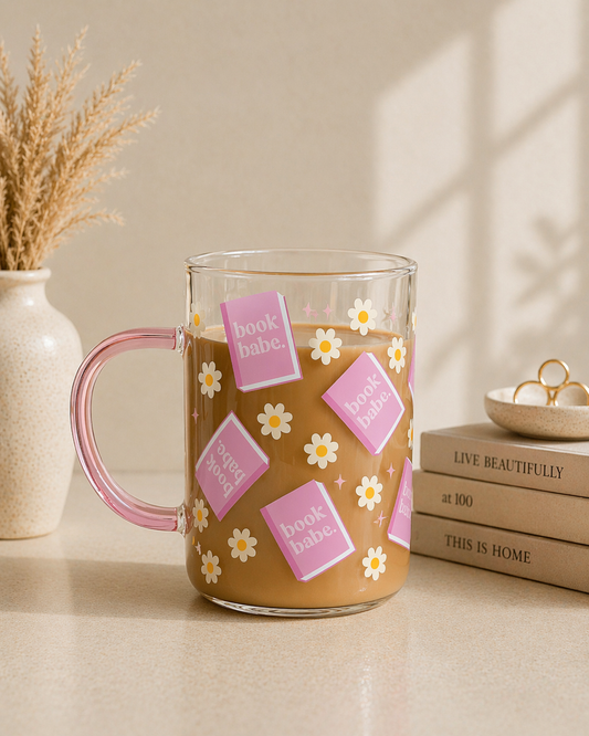 clear glass coffee mug with pink book babe design and daisy pattern being held in hand with books and candle in background