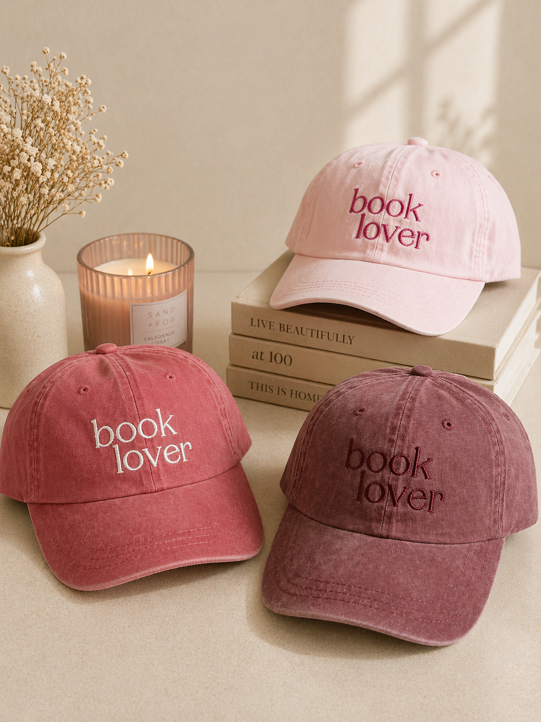 three embroidered book lover hats in pink and red tones styled with books and candle in soft aesthetic setting