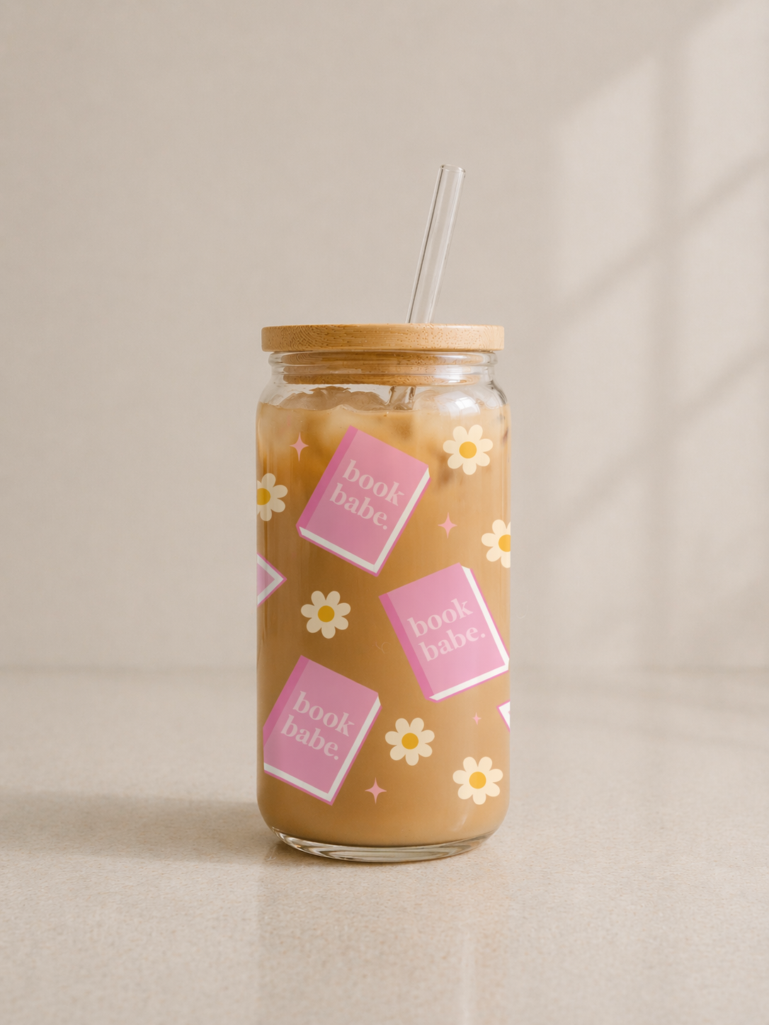 Glass jar with straw lid filled with iced coffee, decorated with 'book babe' and flower designs on a neutral background.
