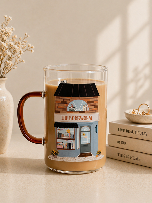 clear glass coffee mug with illustrated bookshop storefront design being held in hand with books in background