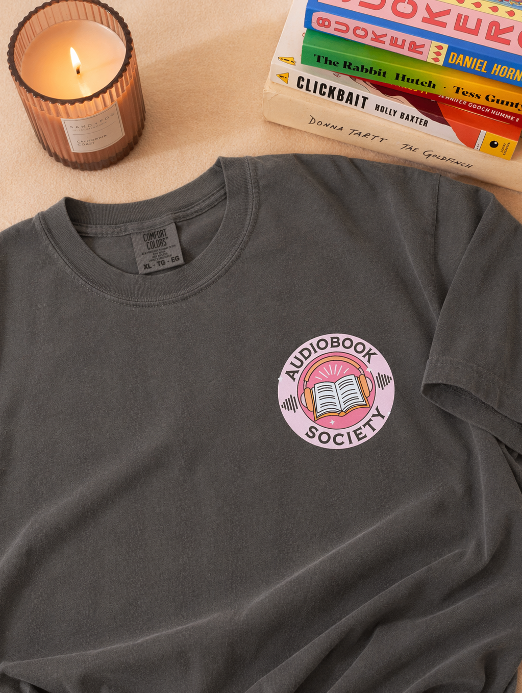 Gray t-shirt with small “Audiobook Society” graphic featuring headphones and book, styled with candle and stacked books