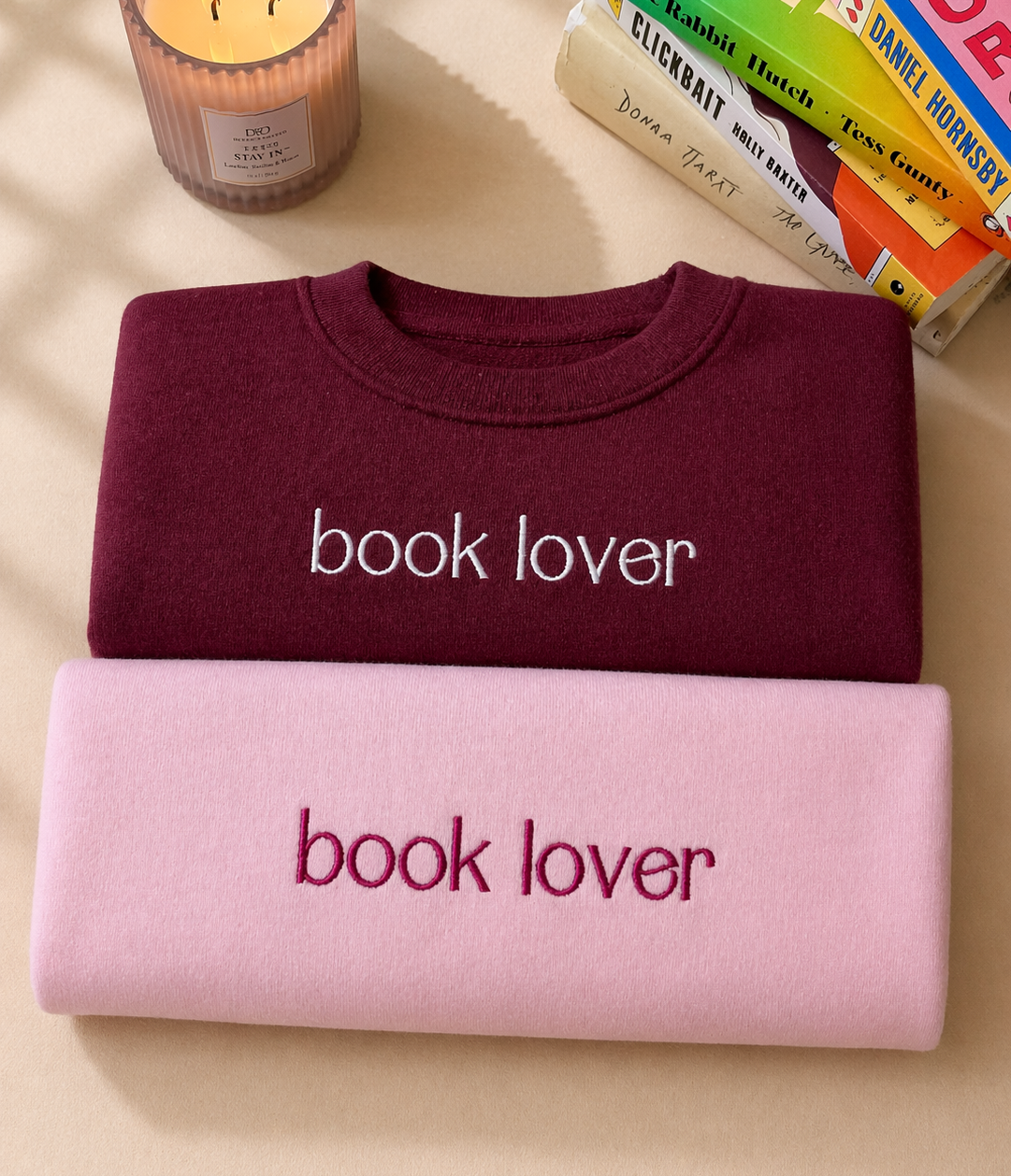 Two sweatshirts, one maroon and one pink, with 'book lover' text on a surface with books and a candle.