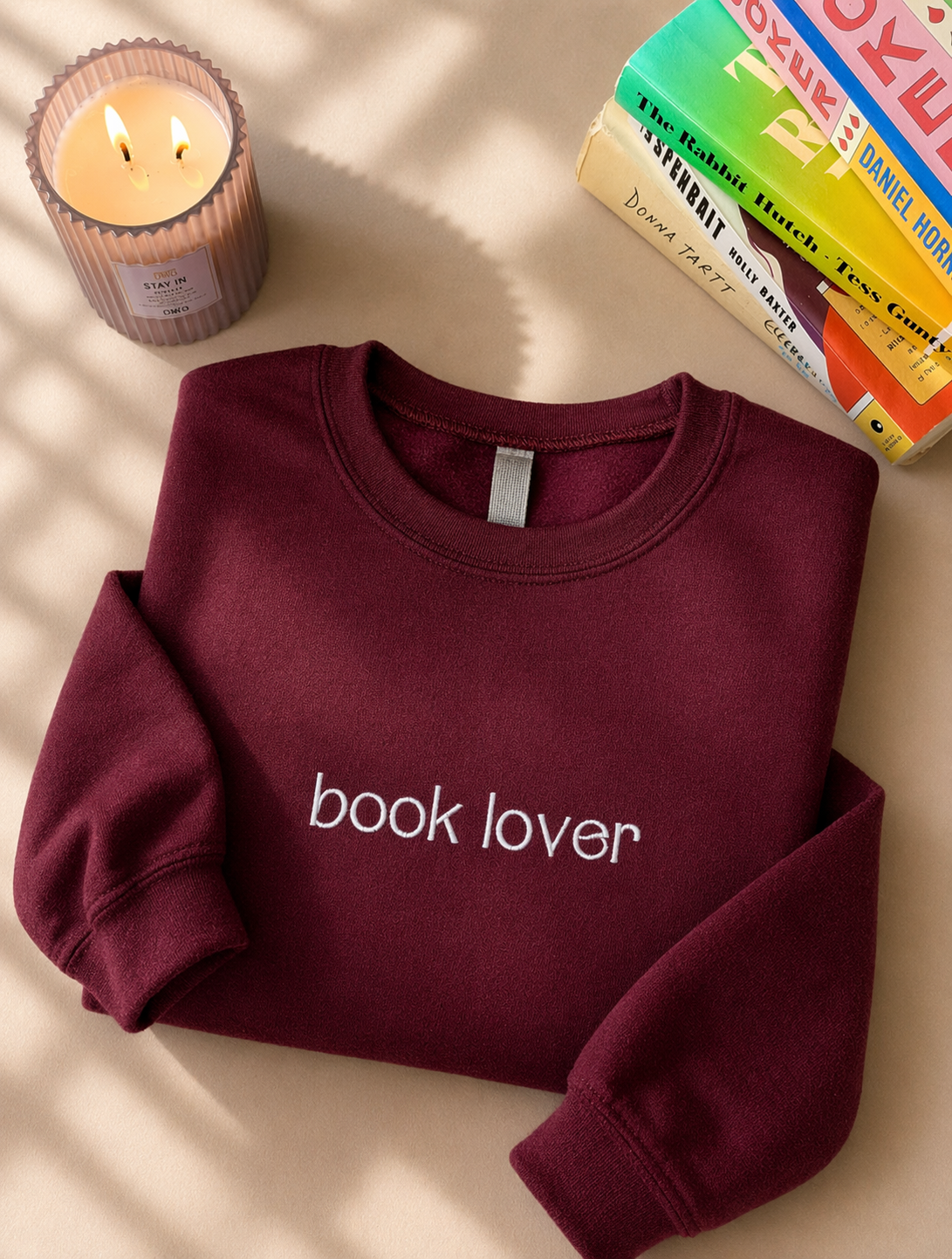 Maroon sweatshirt with 'book lover' text on a beige surface with a candle and books.