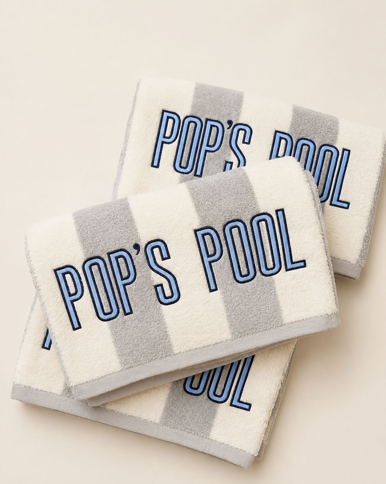 Three striped bath towels stacked on a light beige background, featuring gray and white stripes with 'POP'S POOL' embroidered in blue outlined lettering.