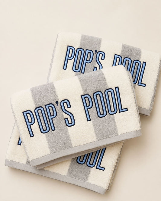 Three striped bath towels stacked on a light beige background, featuring gray and white stripes with 'POP'S POOL' embroidered in blue outlined lettering.