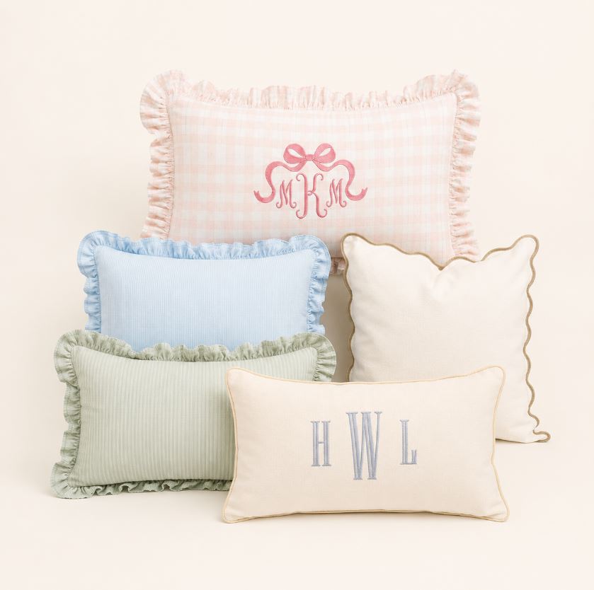 Five decorative throw pillows in soft pastel colors arranged on a light cream background. The pillows include pink gingham, light blue, sage green ribbed, cream with gold trim, and cream with gray monogram letters.