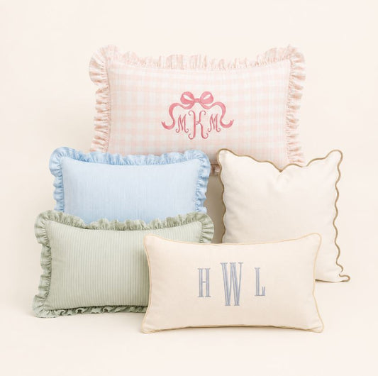 Five decorative throw pillows in soft pastel colors arranged on a light cream background. The pillows include pink gingham, light blue, sage green ribbed, cream with gold trim, and cream with gray monogram letters.