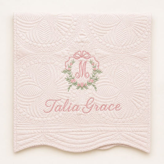 A light pink quilted fabric panel with scalloped edges, featuring an embroidered monogram 'M' in a floral wreath and the name 'Talia Grace' in pink script.