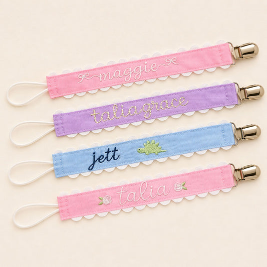 Personalized pacifier clips with embroidered names in pink, lavender, and blue with scalloped trim