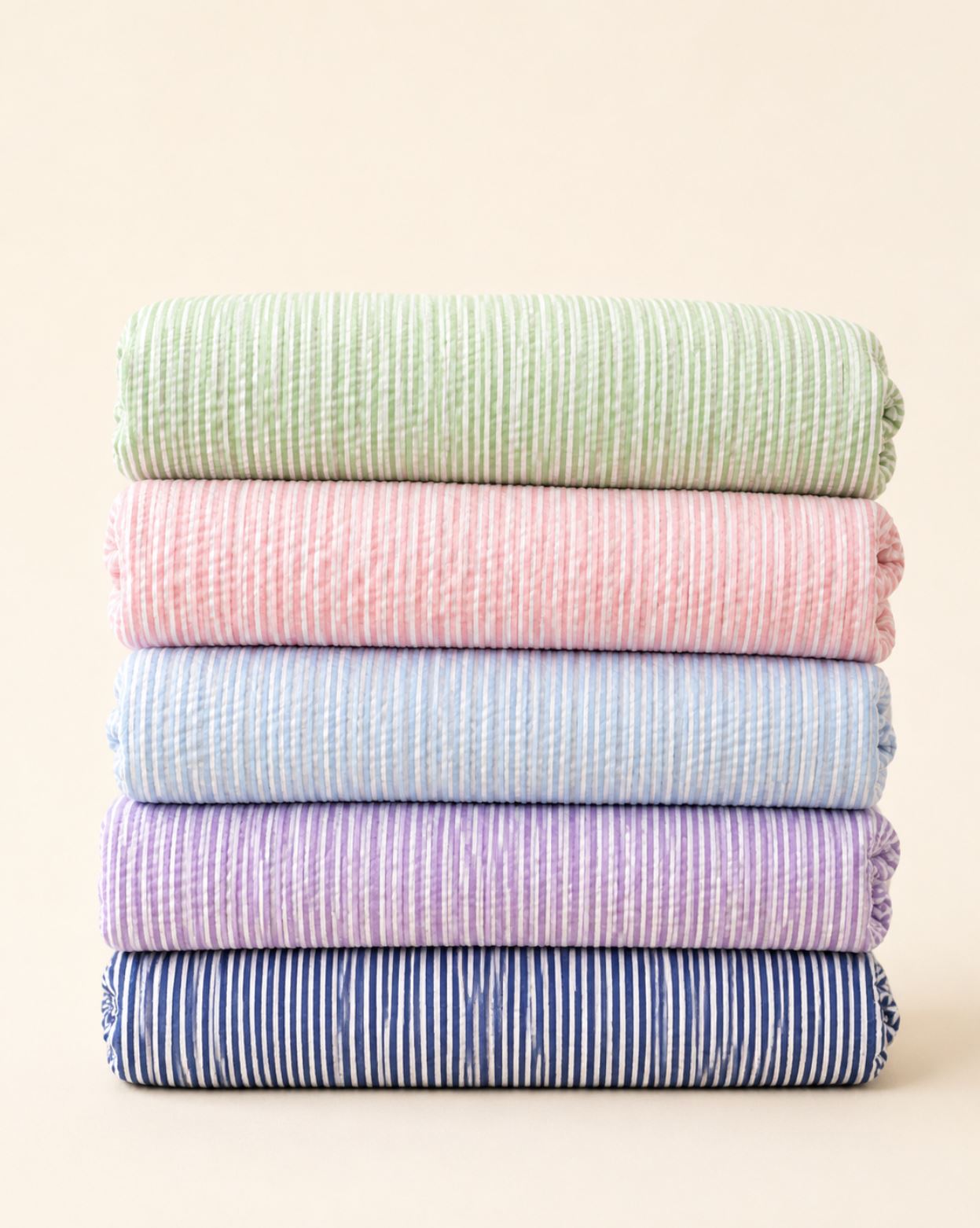 A stack of five folded textured cotton muslin cloths in pastel colors, including sage green, pink, light blue, lavender, and navy blue with white stripes, set against a solid cream background.