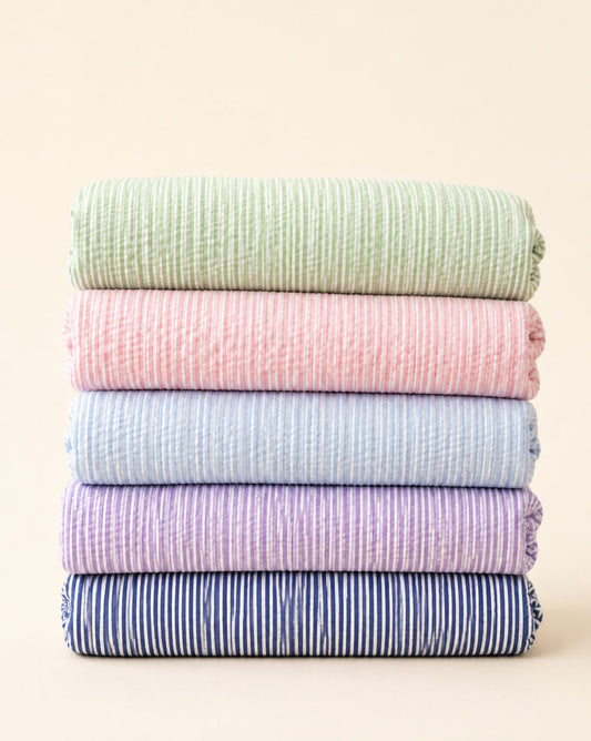 A stack of five folded textured cotton muslin cloths in pastel colors, including sage green, pink, light blue, lavender, and navy blue with white stripes, set against a solid cream background.