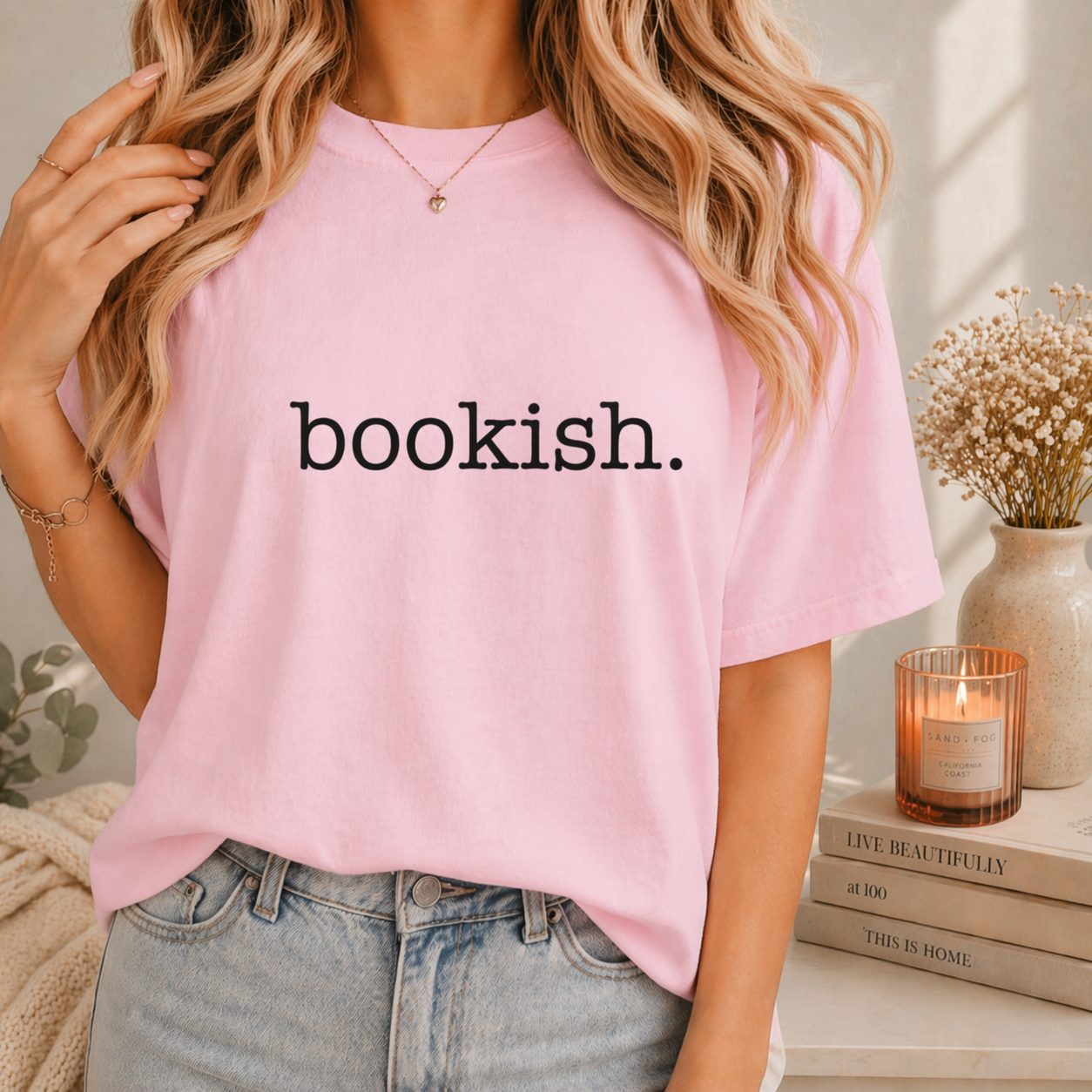 Person wearing a pink t-shirt with 'bookish.' text, standing in a cozy room with books and a candle.