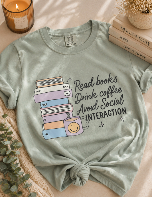 Light green t-shirt with book and coffee graphic and text on a surface with books and a candle.
