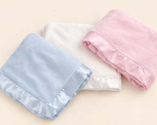 Three folded baby blankets in light blue, white, and soft pink, each with a satin ruffled trim, arranged on a light beige background.