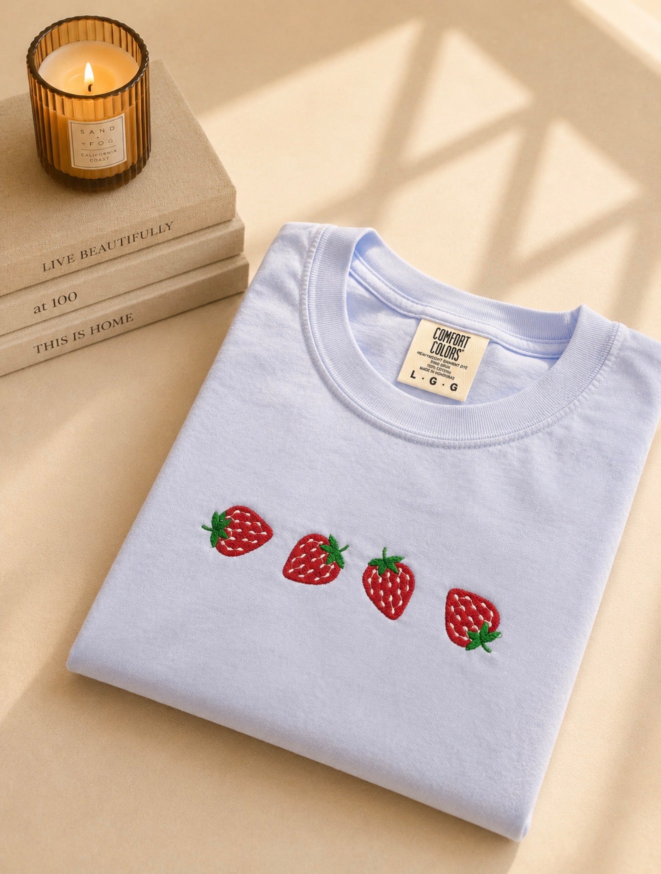 Hydrangea comfort color tee with embroidered strawberries on a beige surface with books and a candle.