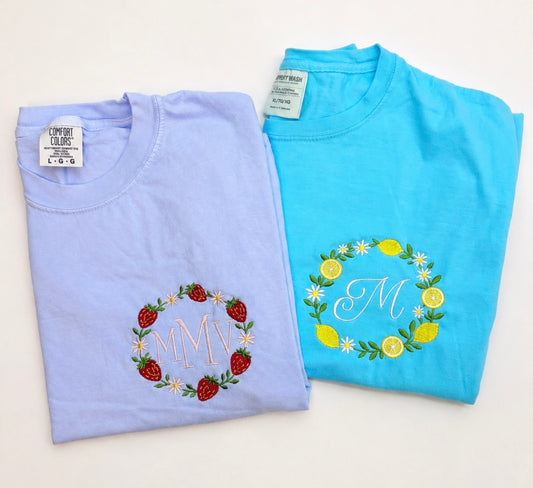 Flat lay of personalized embroidered Comfort Colors t-shirts featuring strawberry and lemon wreath designs with custom monograms embroidered in the center.